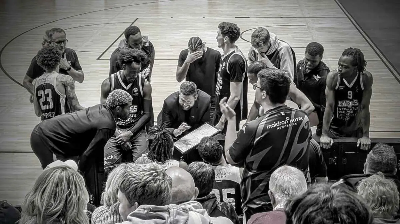 Black and white photograph of Marc Steutel, Head Coach of Newcastle Eagles basketball team, coaching his players with the use of a whiteboard.