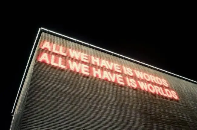 Auto-generated description: A large illuminated sign on a building reads ALL WE HAVE IS WORDS ALL WE HAVE IS WORLDS.
