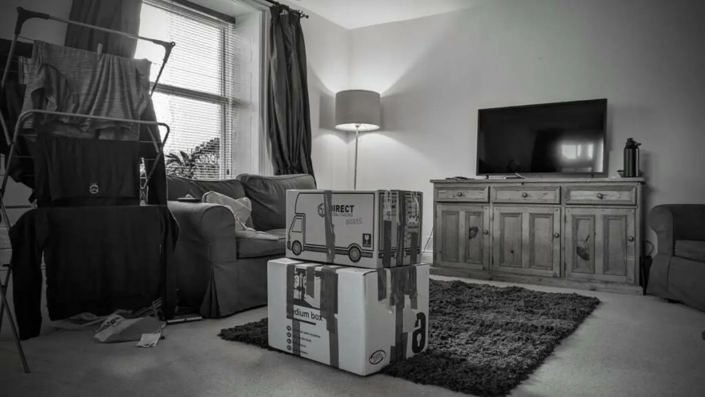 A monochrome photo of a living room with a clothes drying rack, packed moving boxes in the centre, a television on a wooden cabinet, and a cozy armchair, indicating a moving scenario.