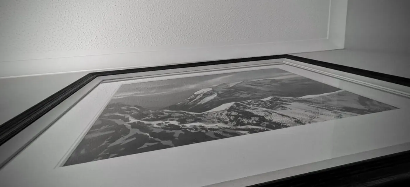 Looking upwards at a narrow angle at a painting of snowy mountains in a black frame.