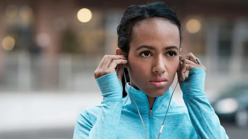 A woman in a blue running top putting in earphones on a city street