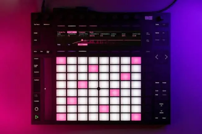 Auto-generated description: A music production controller with a grid of illuminated pads and a display screen is set against a gradient background of pink and purple lighting.