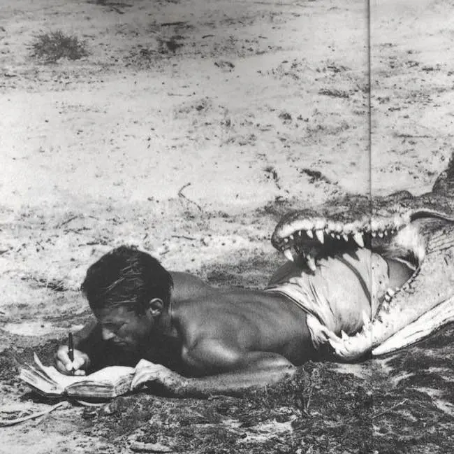 Auto-generated description: A man lies on the ground writing in a notebook while seemingly being swallowed by the open mouth of a large alligator or crocodile.