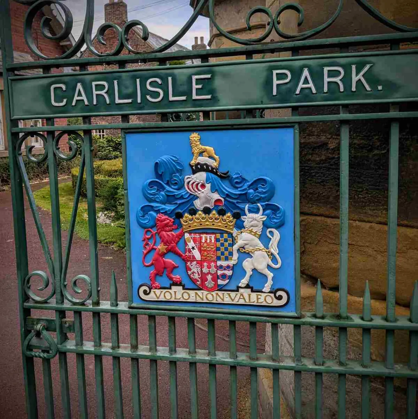Ornate cast-iron gate painted green with words CARLISLE PARK in white lettering. A coat of arms in bold colours is shown with the Latin inscription "VOLO NON VALEO"