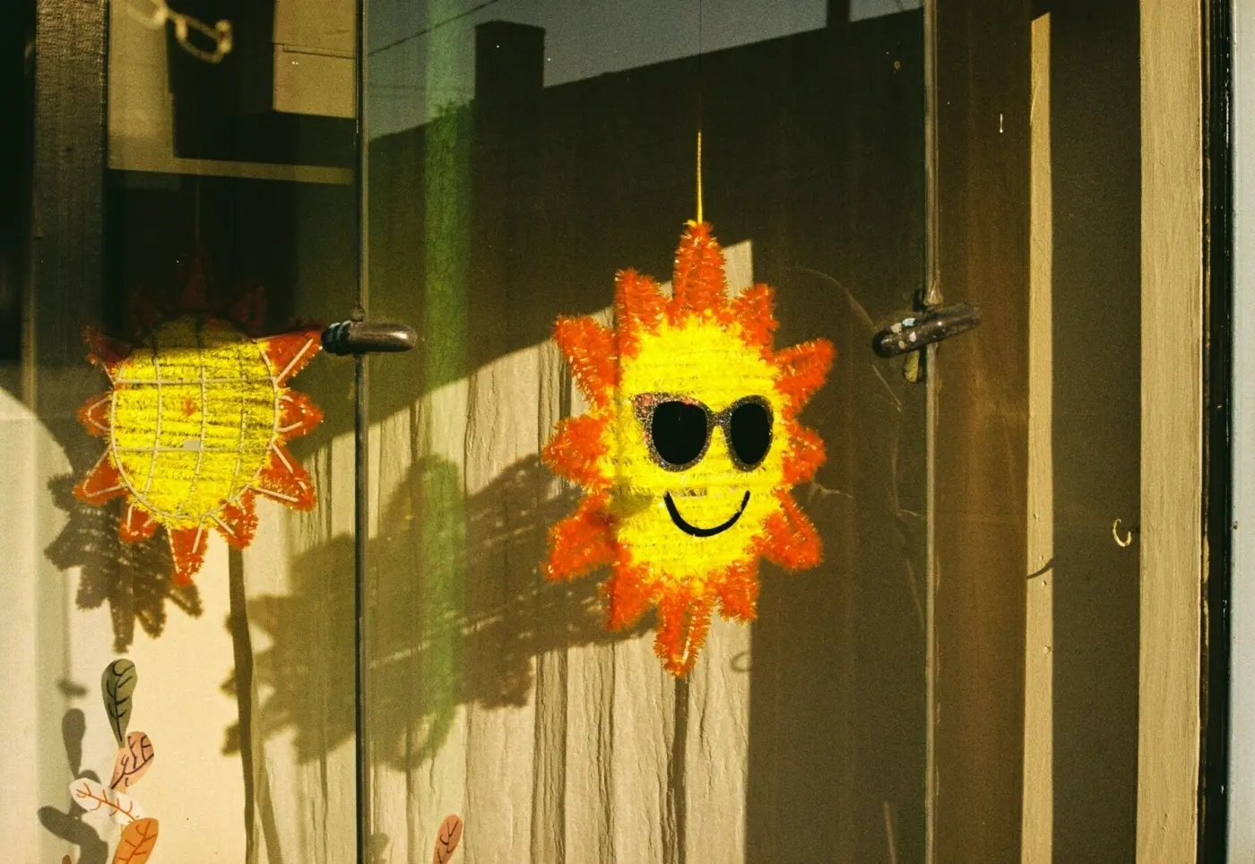 Auto-generated description: A sun-shaped piñata with sunglasses hangs in a window, casting a bright reflection.